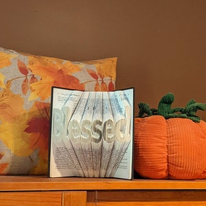 May include: A book folded into the shape of the word "Blessed" with a fall-themed pillow and a plush orange pumpkin on a wooden surface.