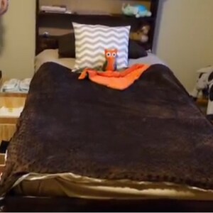 Puede incluir: Una cama marr&oacute;n con una manta marr&oacute;n y una almohada con estampado gris y blanco. Hay un animal de peluche de b&uacute;ho naranja en la cama y un caballo de balanc&iacute;n blanco y negro en el fondo.