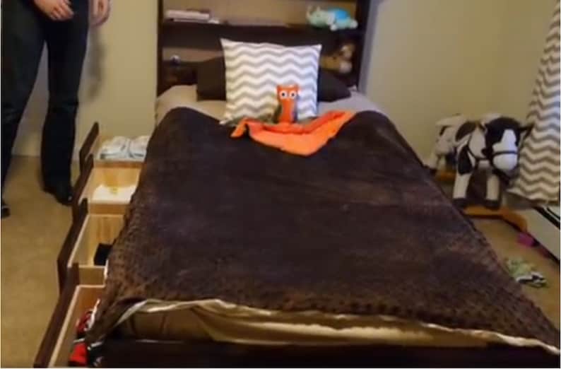 Puede incluir: Una cama marr&oacute;n con una manta marr&oacute;n y una almohada con estampado gris y blanco. Hay un animal de peluche de b&uacute;ho naranja en la cama y un caballo de balanc&iacute;n blanco y negro en el fondo.
