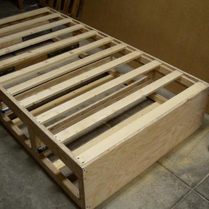 Puede incluir: Un marco de cama de madera con listones y un fondo de madera contrachapada. El marco est&aacute; sin terminar y tiene un color de madera natural.