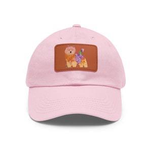 May include: A pale pink baseball cap with a curved peak. It has a brown leather patch with a colourful illustration of a dog made of flowers. The dog has a pink head, orange body, and purple flowers.