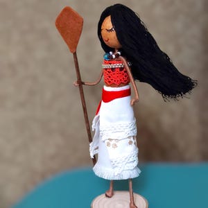 Pu&ograve; includere: Una bambola fatta a mano con lunghi capelli neri, vestita con un abito rosso e bianco, che tiene una pagaia di legno. La bambola &egrave; in piedi su una base di legno.