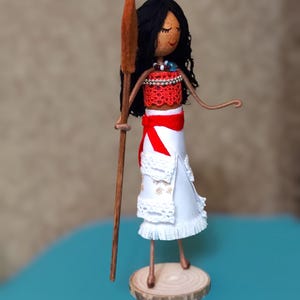 Pu&ograve; includere: Una bambola fatta a mano con i capelli castani, vestita con un abito rosso e bianco, che tiene una pagaia di legno. La bambola &egrave; in piedi su una base di legno.