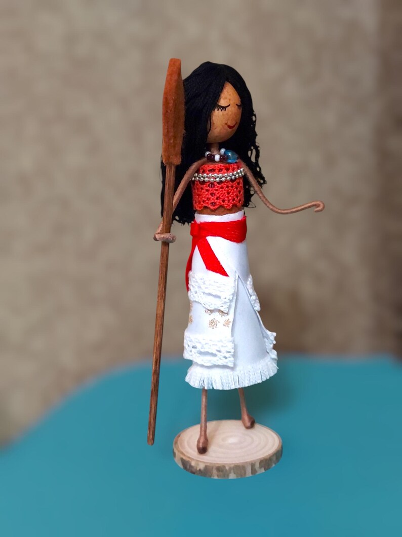 Pu&ograve; includere: Una bambola fatta a mano con i capelli castani, vestita con un abito rosso e bianco, che tiene una pagaia di legno. La bambola &egrave; in piedi su una base di legno.
