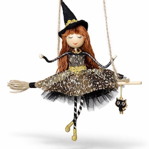 May include: A whimsical Halloween witch doll ornament. The doll has red hair, a black hat, and a leopard print dress. It is suspended from a wooden broom and a black cat charm hangs below. The doll's legs are striped black and white.