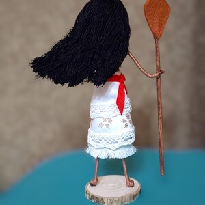 Pu&ograve; includere: Una bambola fatta a mano con lunghi capelli neri, vestita con un abito bianco con pizzo e una fascia rossa. La bambola tiene un bastone di legno con una cima di argilla marrone.