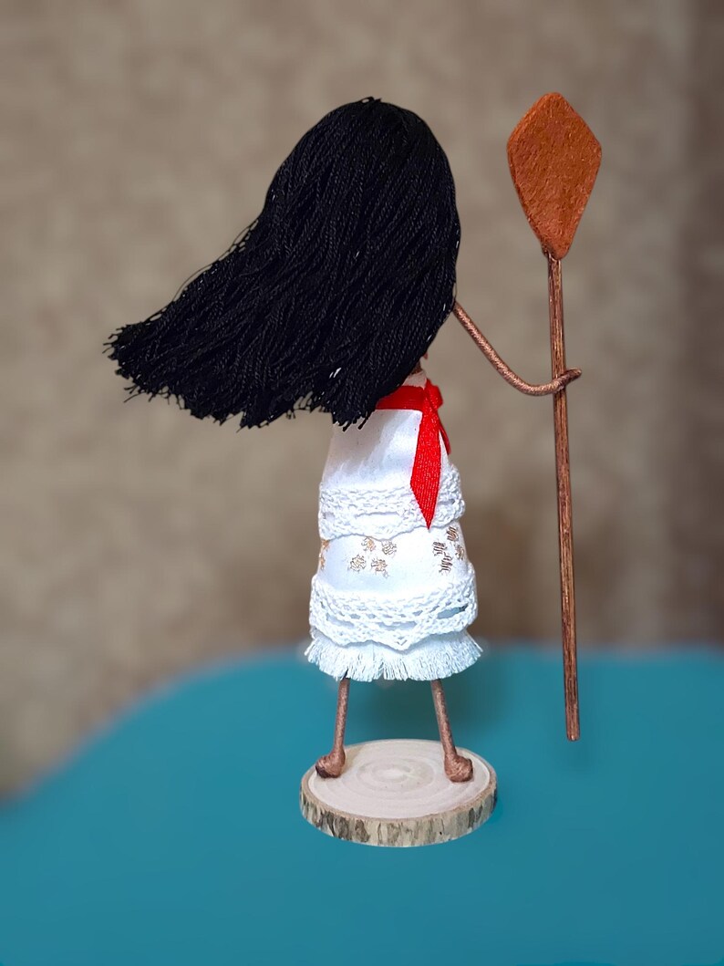 Pu&ograve; includere: Una bambola fatta a mano con lunghi capelli neri, vestita con un abito bianco con pizzo e una fascia rossa. La bambola tiene un bastone di legno con una cima di argilla marrone.