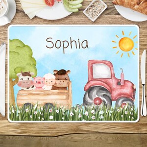 May include: A personalized placemat with a blue background featuring a red tractor, a green tree, a sun, and a wooden wagon with farm animals. The text "Sophia" is printed on the placemat.