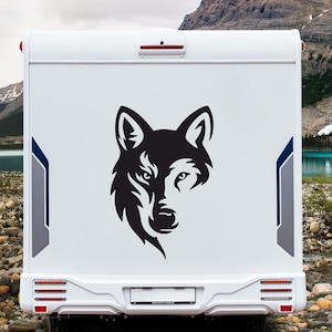 Op de afbeelding: Een witte camper met een zwarte wolfskopsticker op de achterkant. De wolf is afgebeeld in een gestileerd, half gezichtsontwerp. De achtergrond bevat een bergketen, een meer en een bewolkte lucht.