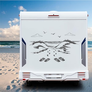 Op de afbeelding: Witte camper met een strandthema-sticker. De sticker toont een grijze illustratie van een strandscène met duinen, voetstappen, slippers en zeemeeuwen. De camper heeft blauwe en grijze accenten aan de zijkanten en staat op een zandstrand.