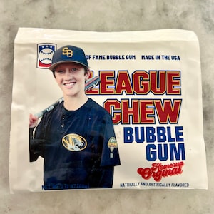 May include: A package of League Chew bubble gum with a baseball player on the front. The package is white with red, blue, and yellow text. The text says "League Chew Bubble Gum" and "Home Run Original".