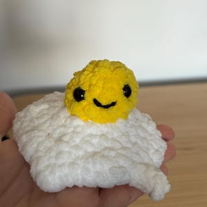May include: A crocheted plush toy shaped like a fried egg, featuring a bright yellow circular yolk with a stitched black smile and two black plastic eyes, centred on a textured, irregular white crocheted egg white base made of soft yarn.
