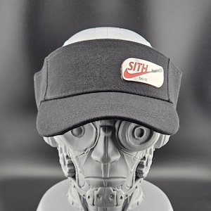May include: Black visor with a white and red patch that reads "SITH" with the Nike swoosh and "DO IT." The visor is displayed on a gray, robotic head. The visor is made of a textured fabric.