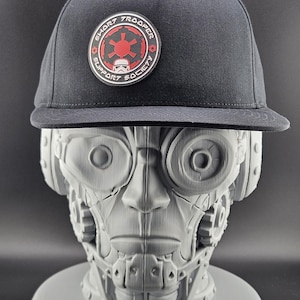 May include: Black flat-brimmed cap with a circular patch featuring the text "SHORT TROOPER SUPPORT SOCIETY" and a red and white graphic. The cap is displayed on a gray bust of a robotic figure.