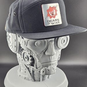 May include: Black flat-brim cap with a rectangular patch featuring a red skull and gear design, and the text "GEARS OF WAR". The cap is displayed on a gray robotic head.