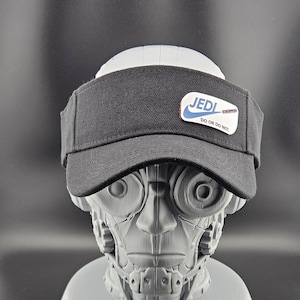 May include: Black visor with a white and blue patch that reads "JEDI DO OR DO NOT". The visor is worn by a grey robotic head. The visor has a curved brim and a black fabric.