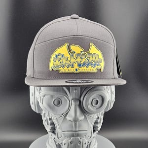 May include: Grey flat-peak cap featuring a yellow and blue graphic with the text "Pocket Monsters" in English and Japanese. The cap has a structured crown and a flat peak. A black tag is attached to the side.