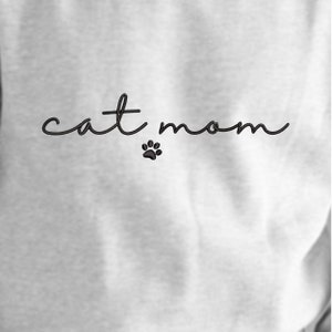 以下が含まれることがあります： 黒い文字で「cat mom」と書いてある白いスウェットシャツ。下に黒い肉球の跡があります。