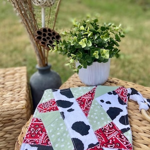 May include: A patchwork baby blanket with a cow print, red bandana print, and light green polka dot fabric. The blanket has a wooden ring attached and is displayed on a woven surface.