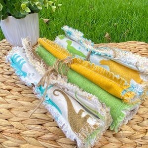 May include: A collection of colorful, rolled fabric cloths tied with twine. The cloths feature dinosaur patterns in shades of green, yellow, and blue. The cloths are on a woven basket, with a green grass background.