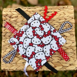 May include: A square baby sensory toy featuring a repeating pattern of Hello Kitty faces with red bows on a white background. The toy has various attached ribbons in red, black, and white patterns, and a red zigzag ribbon.
