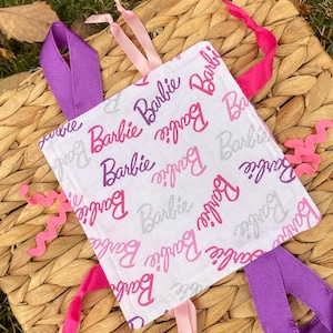 May include: A white square baby tag with the word "Barbie" in pink and silver script. The tag has pink, purple, and light pink ribbons attached to the corners and sides. The tag is on a woven basket.