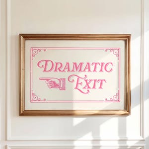 May include: A rectangular wall art print in a gold frame featuring the pink text "DRAMATIC EXIT" in a decorative serif font. To the left of the text is a vintage-style illustration of a pointing hand. The design is enclosed in a pink border.