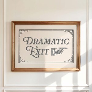 May include: A framed print with the words "Dramatic Exit" in a vintage font. The print features a pointing hand and decorative corner flourishes. The frame is a warm gold color, and the print is mounted on a cream-colored background.