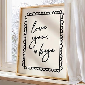 Op de afbeelding: Een ingelijst drukwerk met een witte achtergrond en zwarte tekst die "love you, bye" luidt, met een klein hartje voor het woord "bye". De lijst heeft een decoratieve zwarte rand met gegolfde randen.