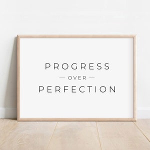 Może przedstawiać: Oprawiony wydruk z napisem "PROGRESS OVER PERFECTION" w minimalistycznej czarnej czcionce. Wydruk jest w jasnej drewnianej ramie i jest wyświetlany na białej ścianie. Widoczne są również słowa "arrie design co" i "DIGITAL DOWNLOAD".