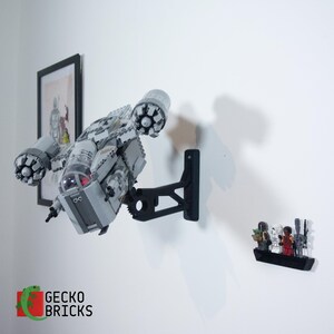May include: A grey and white Lego spaceship model is mounted on a black wall bracket. A framed picture and a small display of Lego figures are also on the white wall. The Gecko Bricks logo is in the lower left corner.
