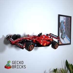 May include: A red Formula 1 race car model, with black tyres and yellow accents, is displayed on a white wall. The model is a detailed replica, showcasing the aerodynamic design of a race car. A framed picture of a race car is on the right.
