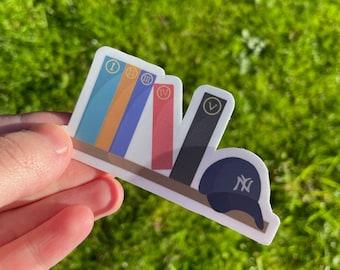 Puede incluir: Una pegatina que representa una estantería con cinco libros de colores numerados con números romanos y una gorra de béisbol azul marino con un logotipo blanco. La pegatina está sobre un fondo de hierba verde.