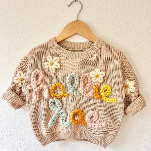 May include: A beige knitted sweater with the word "Hello Rae" embroidered in colorful yarn. The letters are decorated with small white flowers.