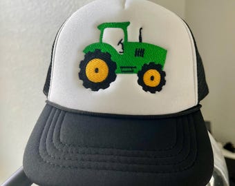May include: A trucker hat with a white front panel featuring a green tractor design with yellow wheels. The hat has a black brim and a black mesh back, with a black rope detail above the brim.