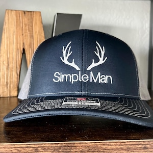 May include: A navy blue trucker hat with a white embroidered antler design and the text "Simple Man." The hat has a gray mesh back and a curved brim. The hat is a size 112.