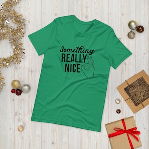May include: A green t-shirt with the phrase "Something Really Nice" in black script, accompanied by a hand gesture. The shirt is laid flat on a light wood surface, with holiday decorations in the background.
