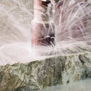 May include: A close-up of a large, rough, gray and green stone being cut by a drill. Water is spraying from the drill bit, creating a mist.
