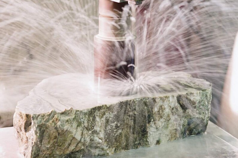 May include: A close-up of a large, rough, gray and green stone being cut by a drill. Water is spraying from the drill bit, creating a mist.
