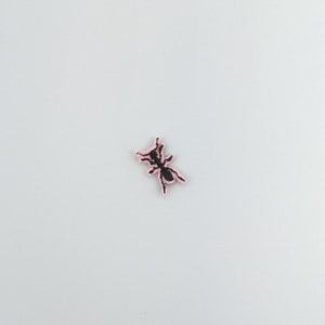 May include: An embroidered patch featuring a black ant with pink stitching. The ant is facing right and has its legs outstretched.
