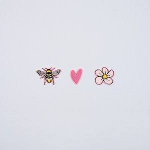 May include: Three embroidered patches on a white background. The patches include a bee with black and yellow details, a pink heart, and a pink flower with a yellow centre and brown outline. These patches are suitable for clothing or accessories.