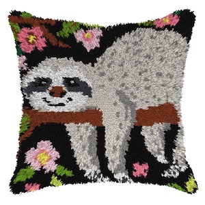 May include: Square cushion with a sloth design. The sloth is grey and white, resting on a brown branch against a black background. Pink flowers and green leaves are scattered around the sloth, adding colour to the design.