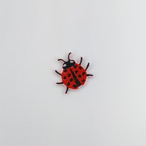 May include: A red ladybug with black spots and antennae, embroidered on a white background.