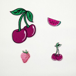 May include: A collection of embroidered fruit patches. The designs include cherries, a watermelon slice, and a strawberry. The patches are in shades of pink, green, and white, with a white border. Ideal for clothing or accessories.