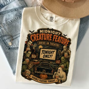 Peut inclure: Un t-shirt couleur crème avec une impression graphique de monstres de films d'horreur classiques dans un ciné-parc. Le texte indique Midnight Creature Feature Drive-In Theatre Tonight Only. Le motif inclut un vampire, une momie et un fantôme.