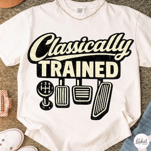 Peut inclure: T-shirt blanc avec l'inscription "Classically Trained" au-dessus d'un dessin d'un levier de vitesse et de pédales de voiture. Le motif est en noir et crème. Le t-shirt est présenté sur une surface claire.