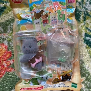 May include: A Sylvanian Families Baby Adventure Series blind bag containing a small, fuzzy gray kitten figurine wearing a pink and brown outfit, encased in clear plastic packaging with a colorful cardboard header featuring various animal characters.