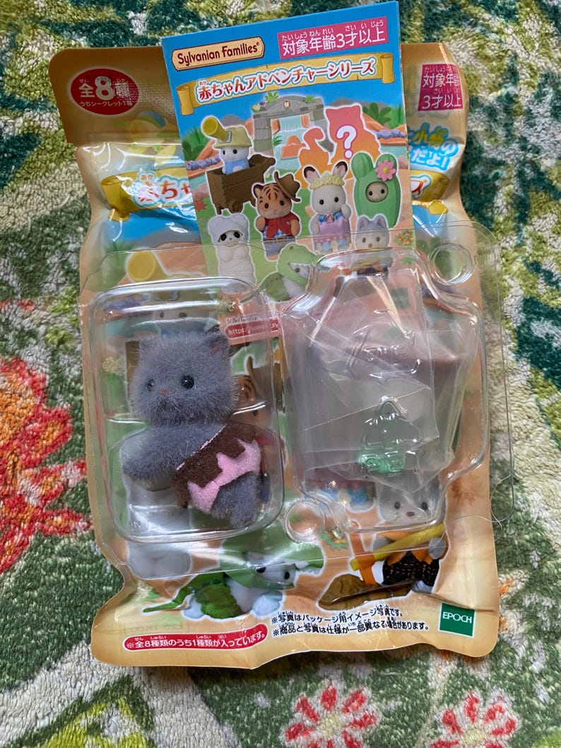 May include: A Sylvanian Families Baby Adventure Series blind bag containing a small, fuzzy gray kitten figurine wearing a pink and brown outfit, encased in clear plastic packaging with a colorful cardboard header featuring various animal characters.