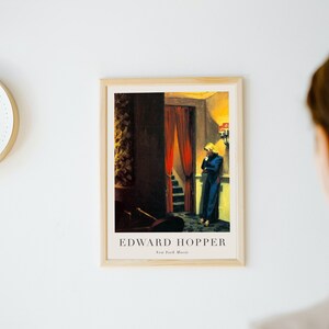 May include: A framed print of a painting by Edward Hopper titled "New York Movie". The painting depicts a woman in a blue dress standing in a dimly lit hallway with a red curtain in the background.