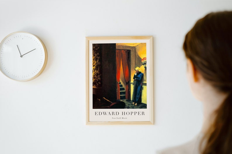 May include: A framed print of a painting by Edward Hopper titled "New York Movie". The painting depicts a woman in a blue dress standing in a dimly lit hallway with a red curtain in the background.